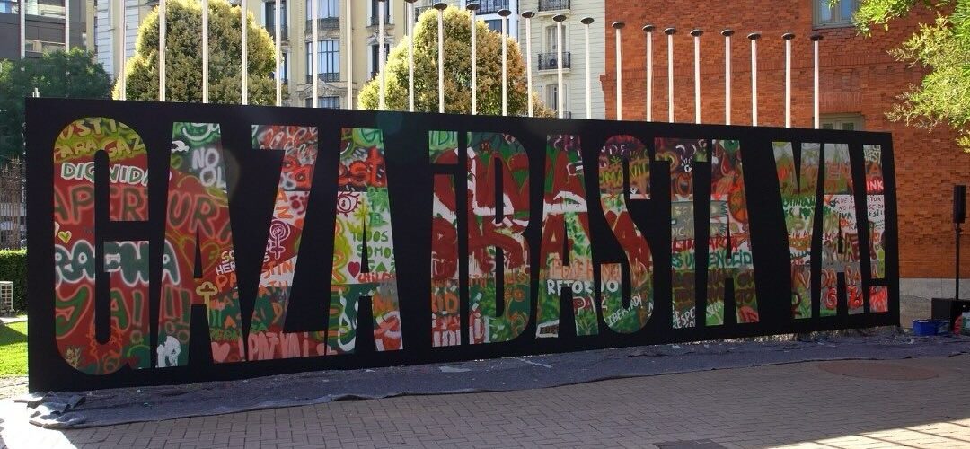 Mural colaborativo facilitado polo colectivo Boa Mistura para a lectura do manifesto na Casa Árabe de Madrid. Sobre un fondo negro, como un caollage de moitas cores e deseños, pódese ler "Gaza basta ya"