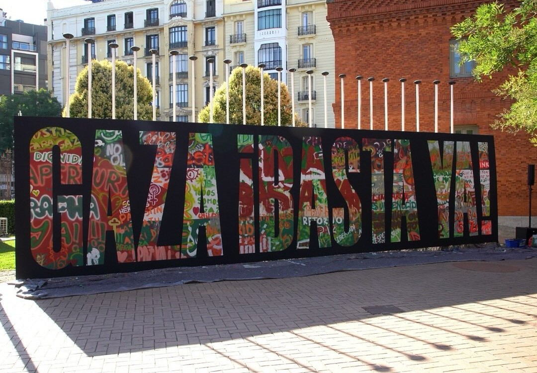 Mural colaborativo facilitado polo colectivo Boa Mistura para a lectura do manifesto na Casa Árabe de Madrid. Sobre un fondo negro, como un caollage de moitas cores e deseños, pódese ler "Gaza basta ya"