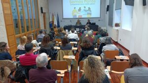 Salón de actos do Edificio Administrativo da Deputación de Ponrevedra cheo de persoas para escoitar a conversa "Cando a cooperación sanda". Na mesa, a deputada de Benestar Social, Paula Bouzós, e a presidenta da Coordinadora Galega de ONGD, María Paz